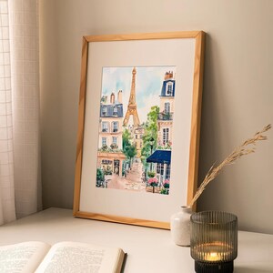 May include: A watercolour painting of a Parisian street scene with the Eiffel Tower in the background. The painting features buildings with balconies, trees, and a cobbled street. The painting is framed in a light wood frame.