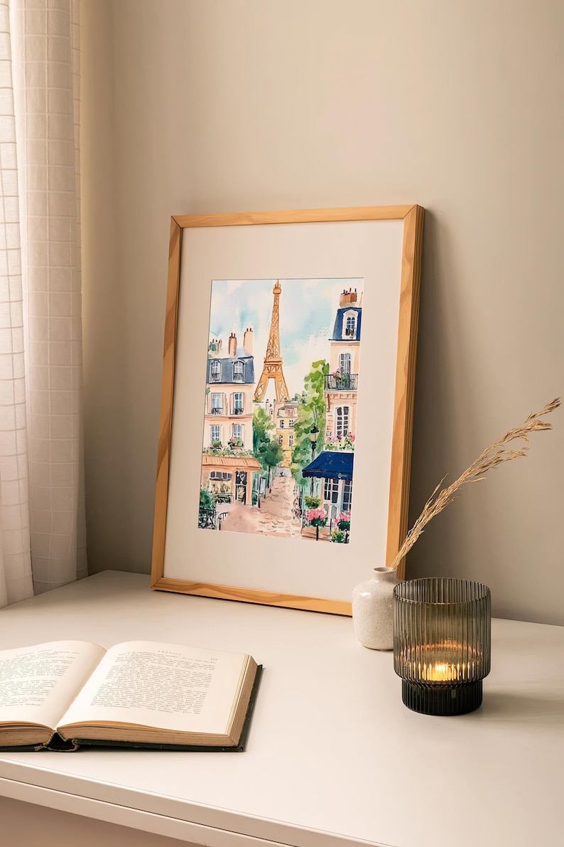 May include: A watercolour painting of a Parisian street scene with the Eiffel Tower in the background. The painting features buildings with balconies, trees, and a cobbled street. The painting is framed in a light wood frame.