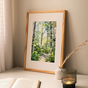 May include: A framed watercolor painting of a forest path with tall trees and green foliage. The painting is on a white table with a book and a candle holder.