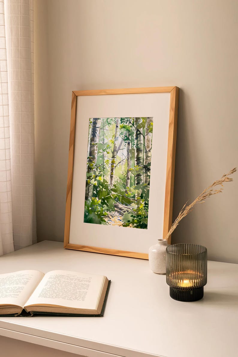 May include: A framed watercolor painting of a forest path with tall trees and green foliage. The painting is on a white table with a book and a candle holder.