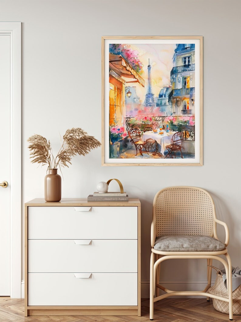 May include: Watercolor painting of a Parisian balcony with a view of the Eiffel Tower. The balcony has a table set for a meal with two chairs. The painting is framed in a light wood frame.