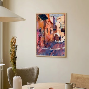 May include: Watercolor painting of a street scene in a wooden frame. The artwork features buildings in shades of orange, pink, and blue, with figures in the foreground. The painting hangs on a beige wall above a round wooden table.