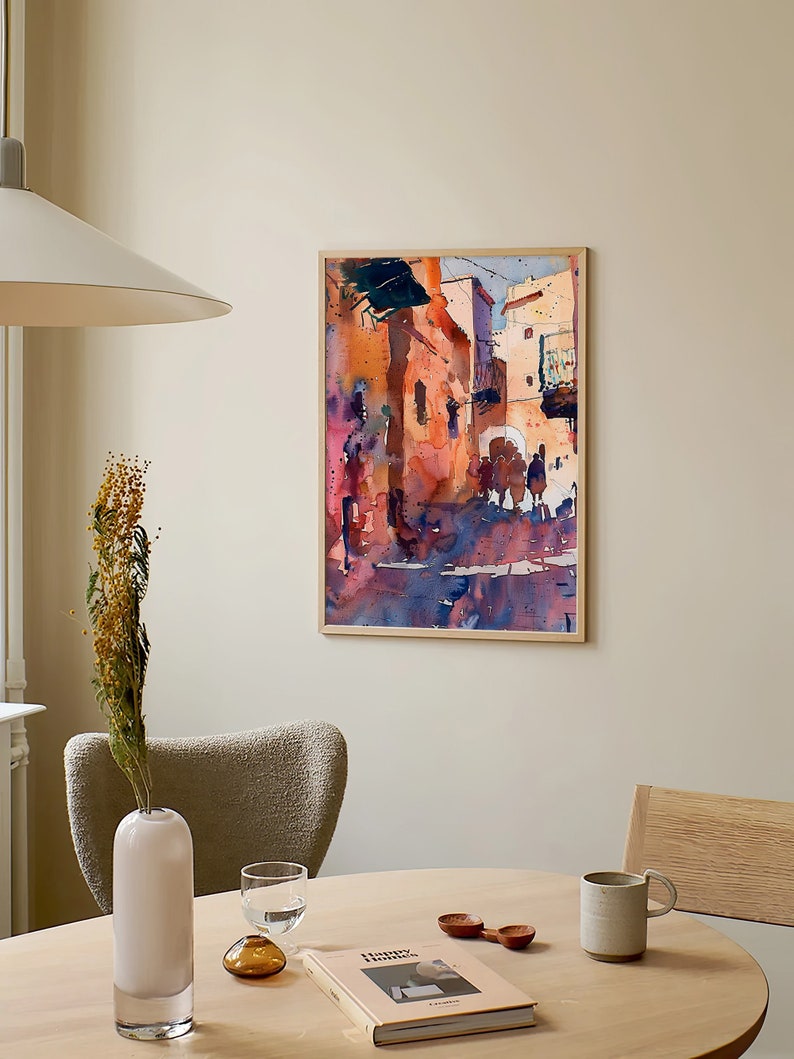 May include: Watercolor painting of a street scene in a wooden frame. The artwork features buildings in shades of orange, pink, and blue, with figures in the foreground. The painting hangs on a beige wall above a round wooden table.