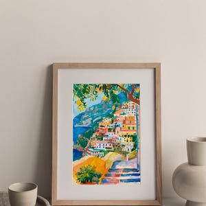 May include: Watercolour painting of a coastal Italian village with colourful buildings cascading down a hillside. The artwork, framed in light wood, features a vibrant palette of blues, yellows, and oranges, capturing the essence of a sunny day.