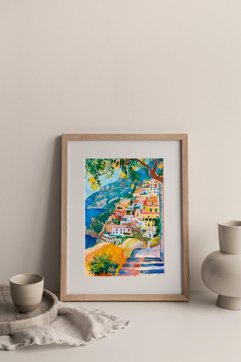 May include: Watercolour painting of a coastal Italian village with colourful buildings cascading down a hillside. The artwork, framed in light wood, features a vibrant palette of blues, yellows, and oranges, capturing the essence of a sunny day.