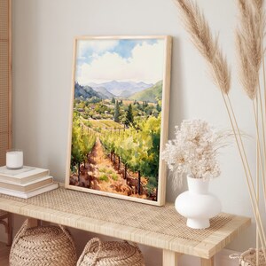 May include: A watercolor painting of a vineyard landscape with rolling hills, green vines, and a small town in the distance. The painting is framed in a light wood frame and is hanging on a wall.