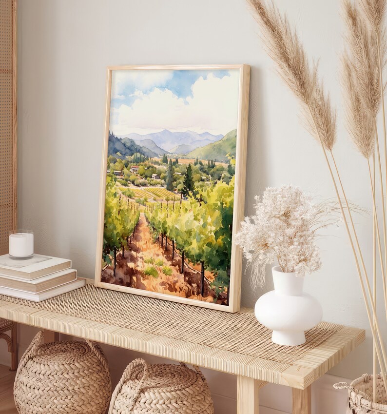 May include: A watercolor painting of a vineyard landscape with rolling hills, green vines, and a small town in the distance. The painting is framed in a light wood frame and is hanging on a wall.