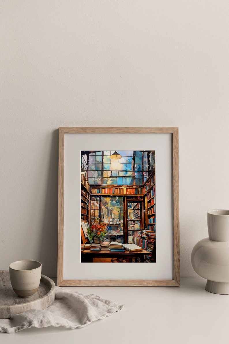 May include: A framed watercolour painting of a bookstore interior with a large window, bookshelves, and a table with books on it. The painting is in a light wood frame and is hanging on a white wall.