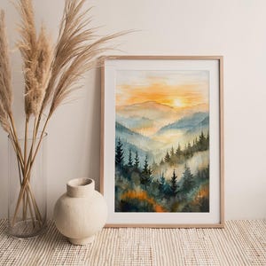 May include: A framed watercolor painting of a mountain landscape at sunset. The artwork features layers of blue and green mountains with a golden sky. The painting is in a light wood frame.
