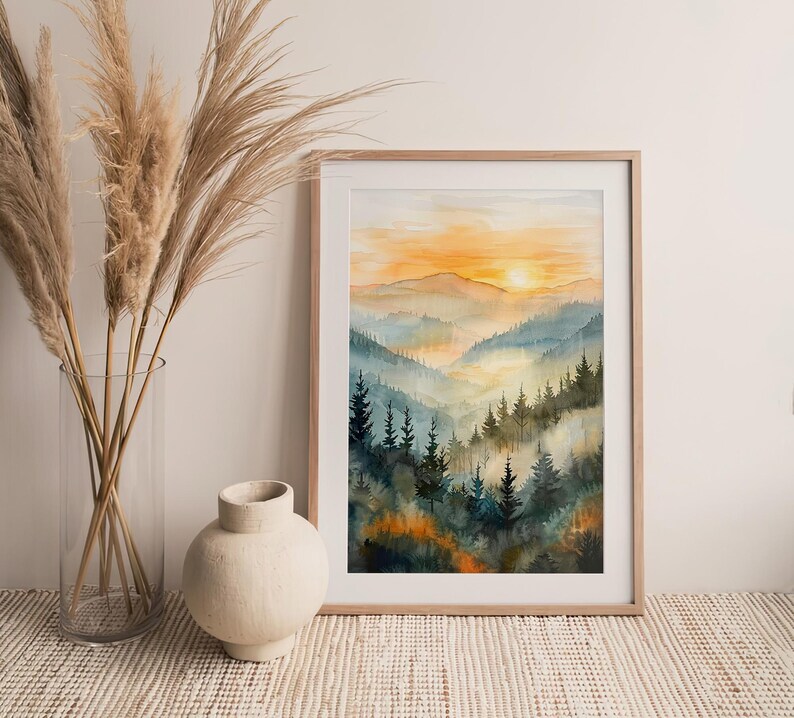 May include: A framed watercolor painting of a mountain landscape at sunset. The artwork features layers of blue and green mountains with a golden sky. The painting is in a light wood frame.
