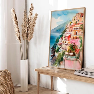 May include: A watercolour painting of a colourful Italian village on a cliffside overlooking the ocean. The painting is framed in a light wood frame and is hanging on a white wall.