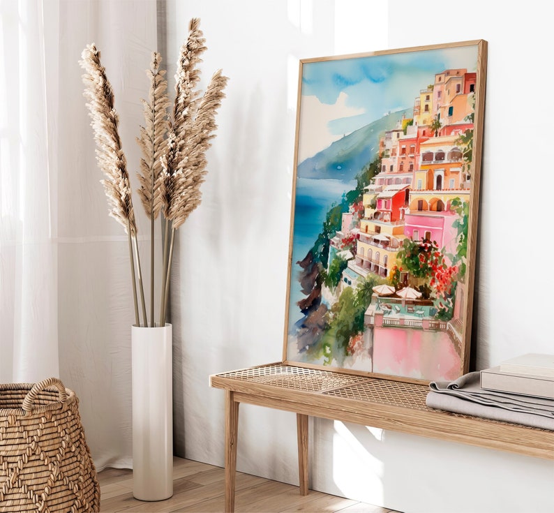May include: A watercolour painting of a colourful Italian village on a cliffside overlooking the ocean. The painting is framed in a light wood frame and is hanging on a white wall.