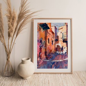 May include: Watercolor painting of a sunlit street scene in a wooden frame. The artwork features buildings in shades of orange, pink, and blue, with silhouetted figures. The painting evokes a warm, Mediterranean atmosphere.