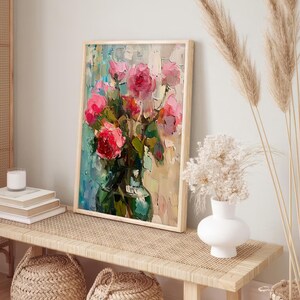 Op de afbeelding: Een ingelijst schilderij van een boeket roze rozen in een groene vaas. Het schilderij staat op een lichte houten tafel met een witte vaas en gedroogde bloemen op de achtergrond.