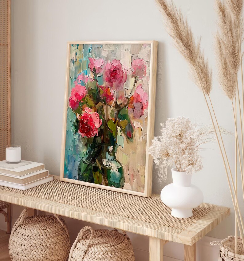 Op de afbeelding: Een ingelijst schilderij van een boeket roze rozen in een groene vaas. Het schilderij staat op een lichte houten tafel met een witte vaas en gedroogde bloemen op de achtergrond.