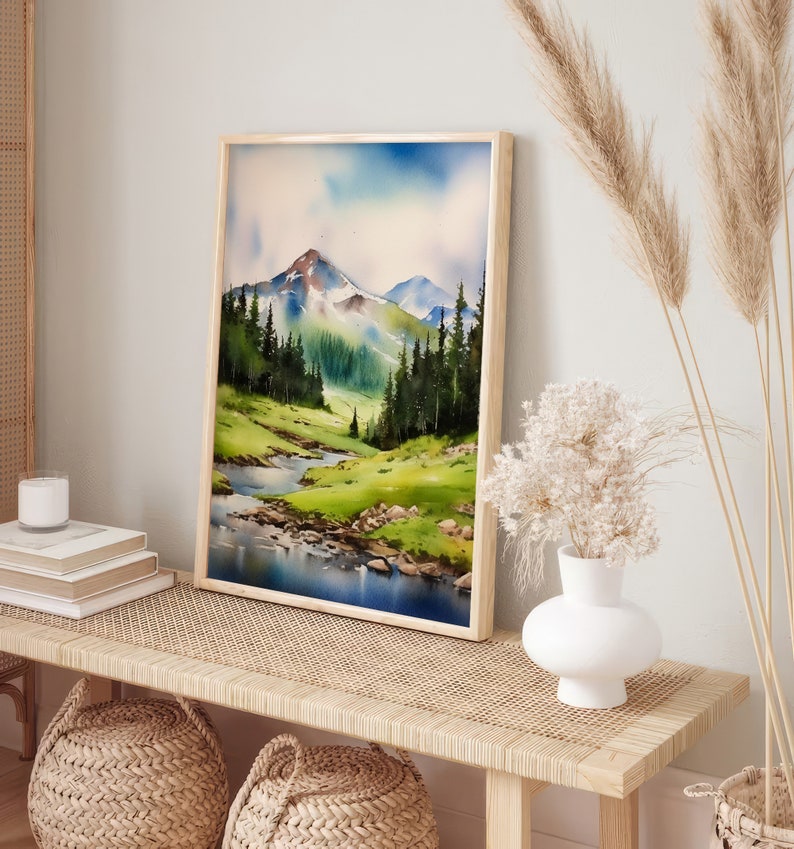 May include: A framed watercolour painting of a mountain landscape with a river flowing through a valley. The painting is in a light wood frame and is hanging on a wall above a wooden bench.