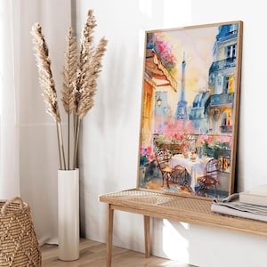 May include: A watercolor painting of a Parisian balcony scene with a view of the Eiffel Tower. The balcony has a table set for a meal with chairs and flowers. The painting is framed in a light wood frame.