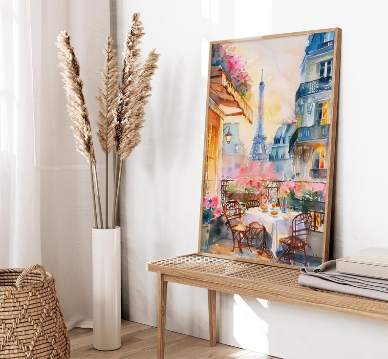 May include: A watercolor painting of a Parisian balcony scene with a view of the Eiffel Tower. The balcony has a table set for a meal with chairs and flowers. The painting is framed in a light wood frame.