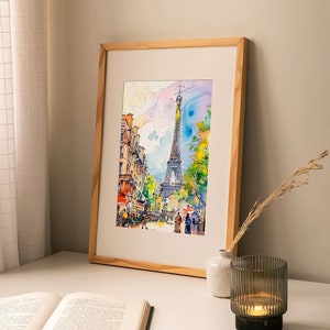 Puede incluir: Una pintura de acuarela enmarcada de la Torre Eiffel en Par&iacute;s, Francia. La pintura representa una escena de calle con edificios, &aacute;rboles y personas caminando. La pintura est&aacute; en un marco de madera clara y se exhibe en una mesa blanca con un libro y un portavelas.
