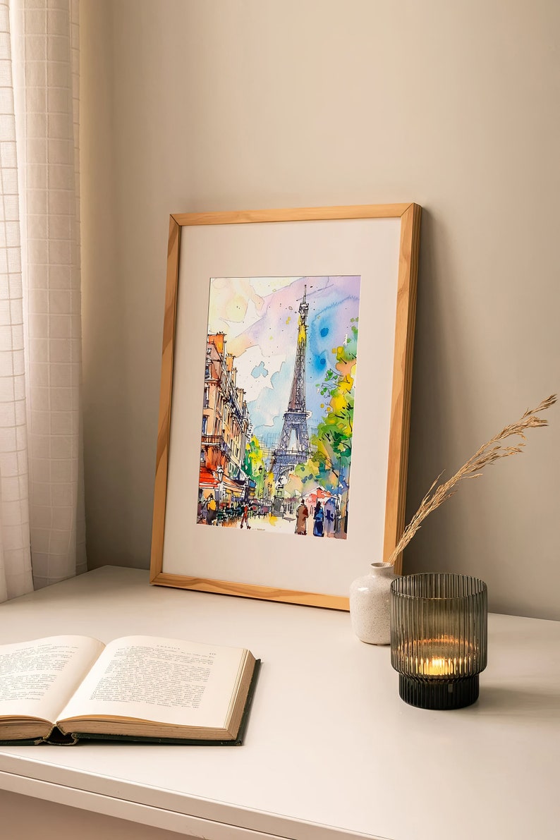 Puede incluir: Una pintura de acuarela enmarcada de la Torre Eiffel en Par&iacute;s, Francia. La pintura representa una escena de calle con edificios, &aacute;rboles y personas caminando. La pintura est&aacute; en un marco de madera clara y se exhibe en una mesa blanca con un libro y un portavelas.