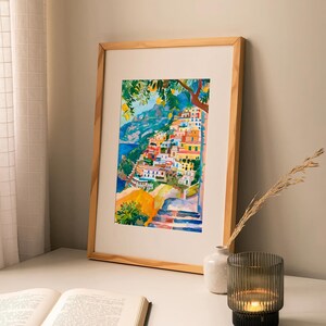 May include: Watercolour painting of a coastal village with colourful buildings, framed in a light wood frame. The artwork depicts a hillside town with blue water and green trees. A book, vase, and candle holder are on the table.