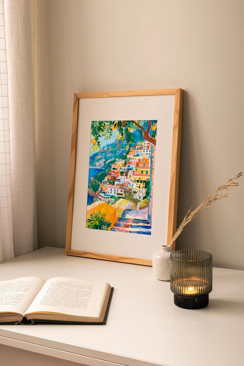May include: Watercolour painting of a coastal village with colourful buildings, framed in a light wood frame. The artwork depicts a hillside town with blue water and green trees. A book, vase, and candle holder are on the table.