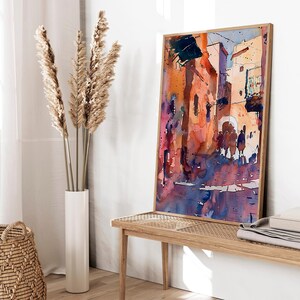 May include: Watercolor painting of a street scene in warm tones. The artwork features buildings in shades of orange, red, and purple, with figures in the street. The painting is framed in a light wood frame and leans against a white wall.