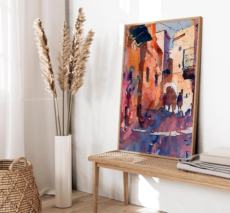 May include: Watercolor painting of a street scene in warm tones. The artwork features buildings in shades of orange, red, and purple, with figures in the street. The painting is framed in a light wood frame and leans against a white wall.