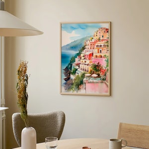 May include: A watercolour painting of a colourful Italian village on a cliff overlooking the sea. The painting is framed in a light wood frame and is hanging on a light beige wall.