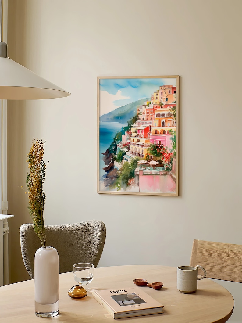 May include: A watercolour painting of a colourful Italian village on a cliff overlooking the sea. The painting is framed in a light wood frame and is hanging on a light beige wall.