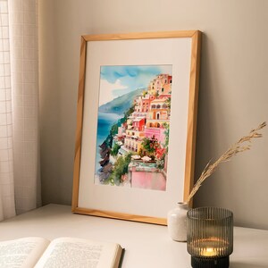 May include: A framed watercolour painting of a colourful Italian village on a cliffside overlooking the sea. The painting is in a light wood frame and is displayed on a white table with a book and a candle holder.