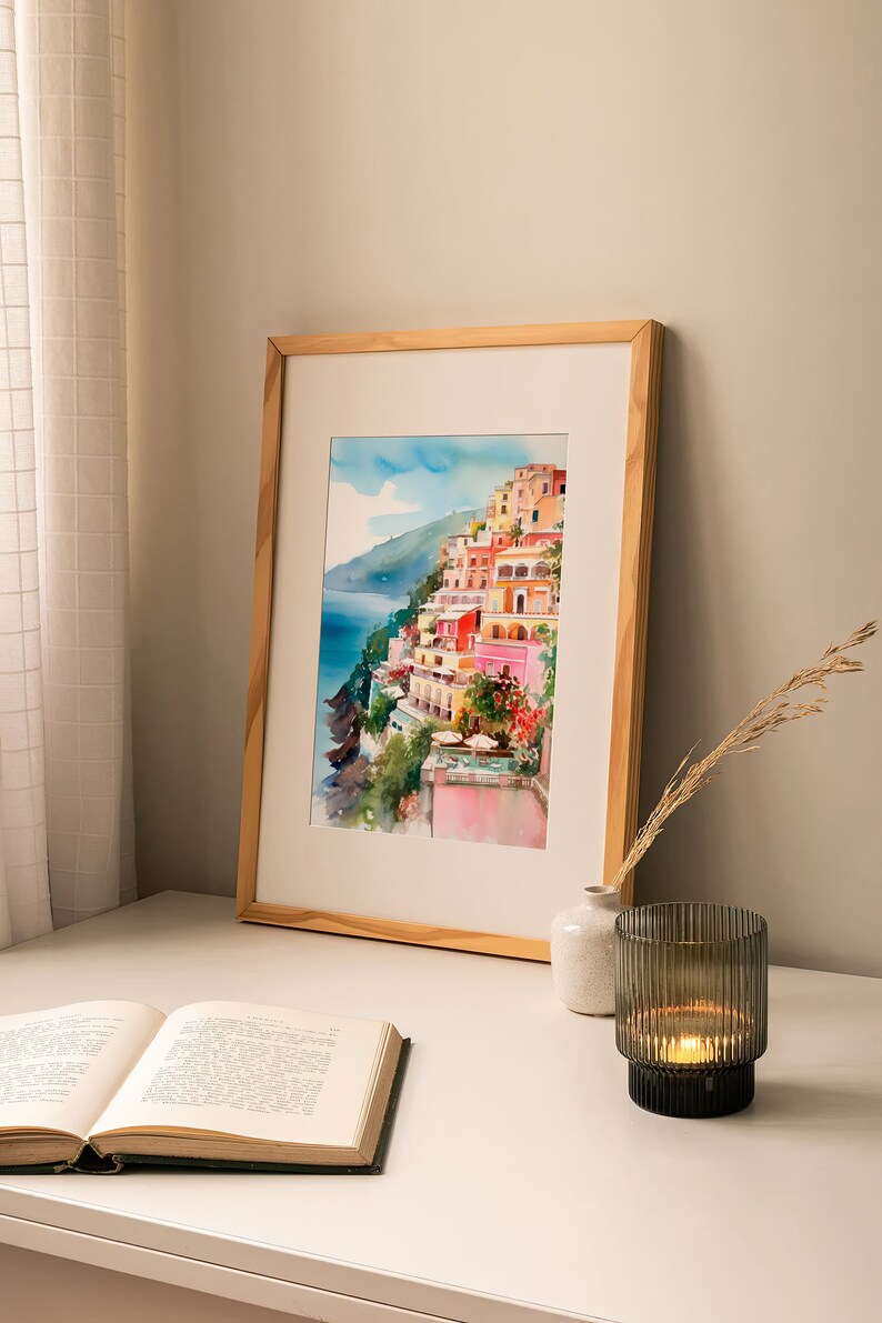 May include: A framed watercolour painting of a colourful Italian village on a cliffside overlooking the sea. The painting is in a light wood frame and is displayed on a white table with a book and a candle holder.