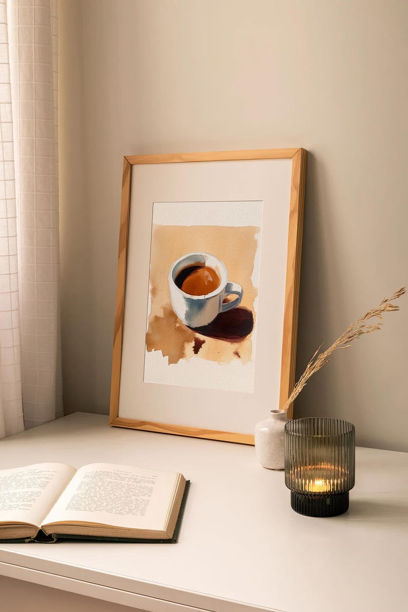 Puede incluir: Una pintura de acuarela enmarcada de una taza de caf&eacute; blanca con un l&iacute;quido marr&oacute;n en su interior. La taza est&aacute; sobre una superficie marr&oacute;n con una mancha marr&oacute;n. La pintura est&aacute; en un marco de madera.
