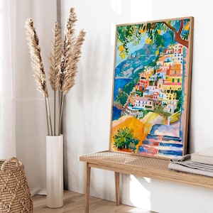 May include: A framed watercolour painting shows a vibrant coastal village with buildings on a hillside. The artwork uses blue, yellow, orange, and red, with a lemon tree in the top right. The painting is displayed on a wooden bench, with a vase of dried plants and a woven basket.