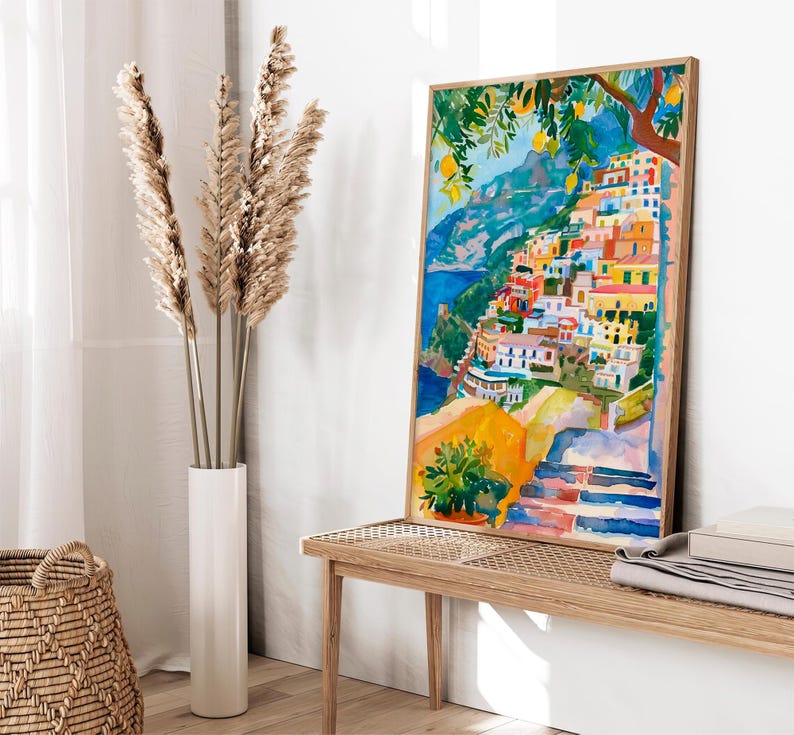 May include: A framed watercolour painting shows a vibrant coastal village with buildings on a hillside. The artwork uses blue, yellow, orange, and red, with a lemon tree in the top right. The painting is displayed on a wooden bench, with a vase of dried plants and a woven basket.