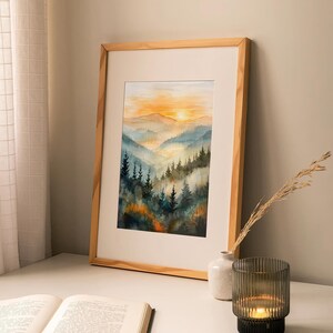 May include: Watercolor painting of a mountain landscape with a sunset. The artwork is framed in a light wood frame and features a vibrant orange and yellow sky, with blue and green trees in the foreground. The painting is displayed on a white surface.