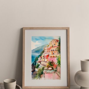 May include: A framed watercolour painting depicting a colourful Italian village perched on a cliff overlooking the sea. The buildings are painted in shades of pink, yellow and orange, with palm trees and lush greenery in the foreground.