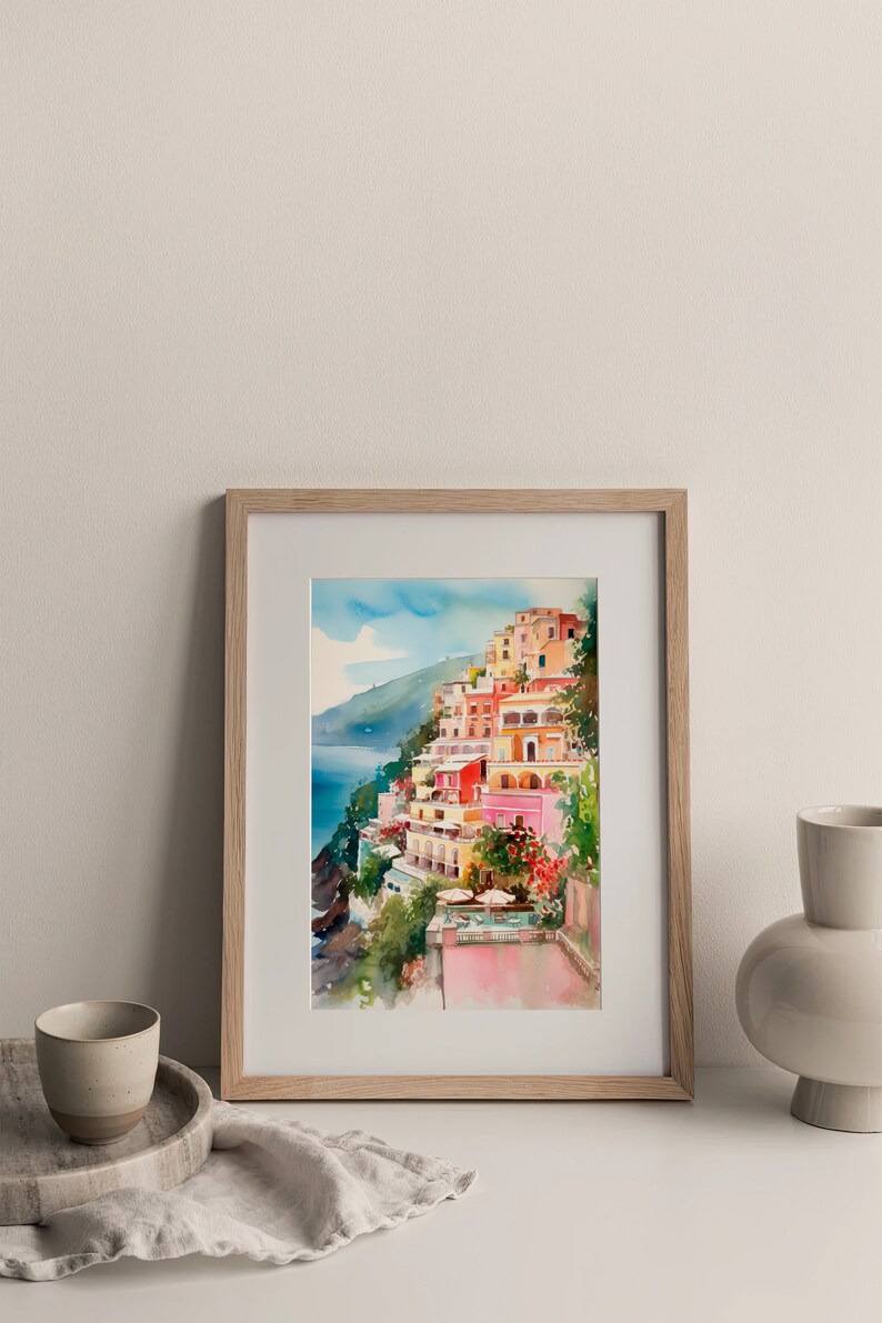 May include: A framed watercolour painting depicting a colourful Italian village perched on a cliff overlooking the sea. The buildings are painted in shades of pink, yellow and orange, with palm trees and lush greenery in the foreground.