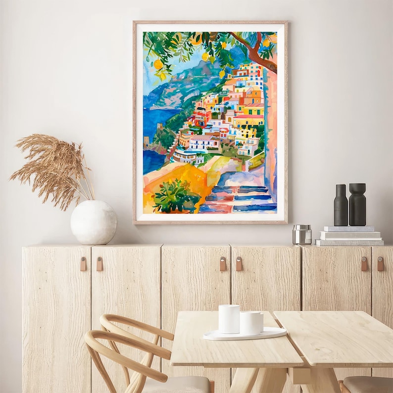 May include: Watercolour painting of a coastal Italian village with colourful buildings, a blue sea, and a tree with lemons. The artwork is framed and displayed on a wooden cabinet. The painting evokes a sense of travel and Mediterranean charm.