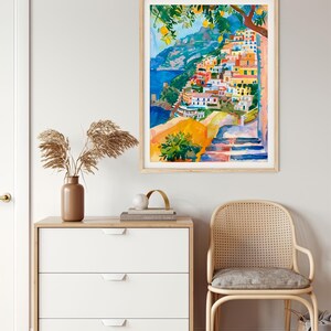 May include: Watercolour painting of a coastal Italian village with colourful buildings on a hillside. The artwork features a blue sea, lemon trees, and a wooden frame. The painting is displayed above a white chest of drawers and a rattan chair.