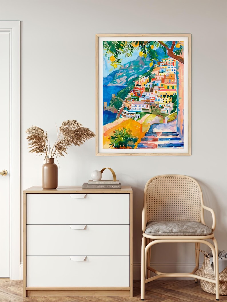 May include: Watercolour painting of a coastal Italian village with colourful buildings on a hillside. The artwork features a blue sea, lemon trees, and a wooden frame. The painting is displayed above a white chest of drawers and a rattan chair.
