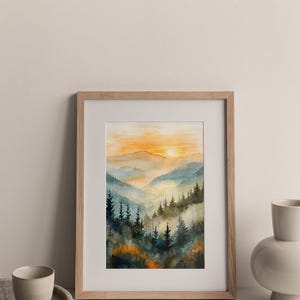 May include: Watercolor painting of a mountain landscape with a sunset. The artwork features layers of blue and green trees, with an orange and yellow sky. The framed art is displayed on a white surface.