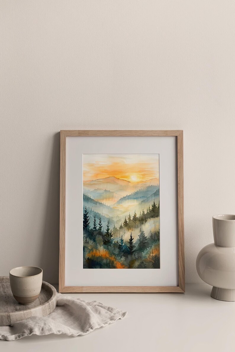 May include: Watercolor painting of a mountain landscape with a sunset. The artwork features layers of blue and green trees, with an orange and yellow sky. The framed art is displayed on a white surface.