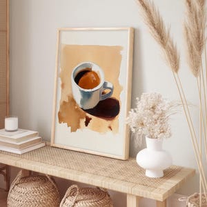 Puede incluir: Una pintura al agua enmarcada de una taza de caf&eacute; blanca con caf&eacute; marr&oacute;n en su interior, sentada sobre una superficie marr&oacute;n con una sombra debajo. La pintura est&aacute; en un estante de madera marr&oacute;n claro con un jarr&oacute;n blanco y flores secas en el lado derecho del estante.