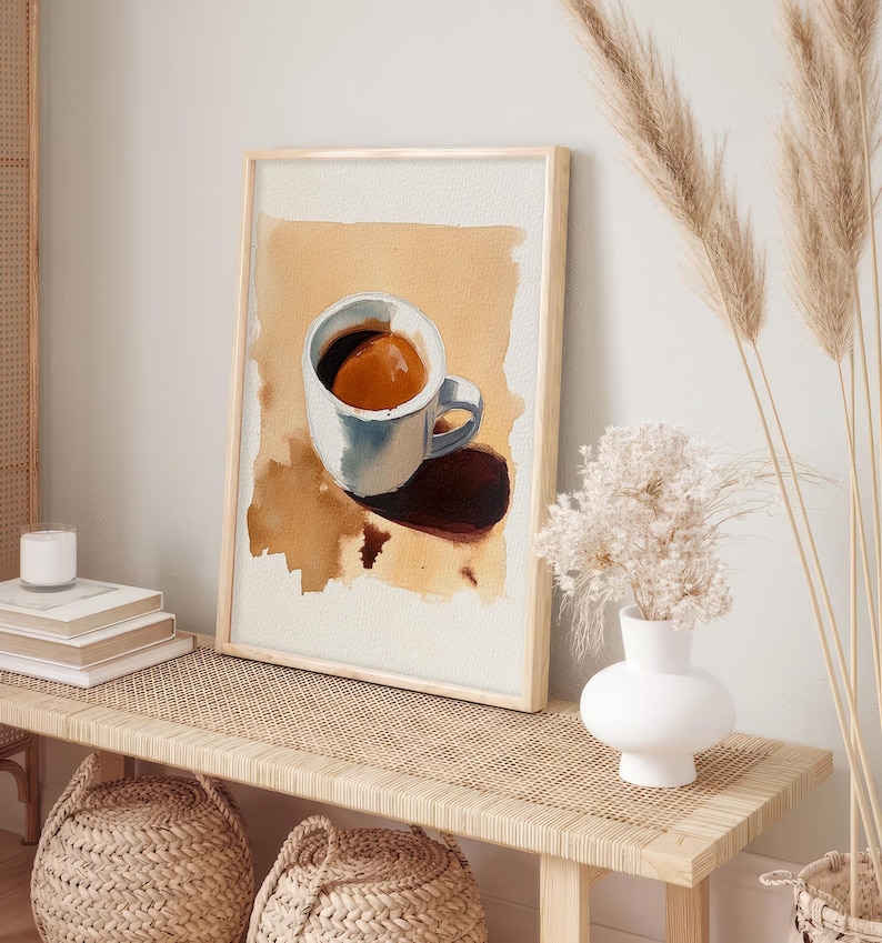 Puede incluir: Una pintura al agua enmarcada de una taza de caf&eacute; blanca con caf&eacute; marr&oacute;n en su interior, sentada sobre una superficie marr&oacute;n con una sombra debajo. La pintura est&aacute; en un estante de madera marr&oacute;n claro con un jarr&oacute;n blanco y flores secas en el lado derecho del estante.