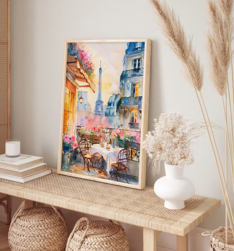 May include: A watercolor painting of a Parisian balcony scene with a view of the Eiffel Tower. The balcony is decorated with flowers and there is a table set for a meal with two chairs. The painting is framed in a light wood frame.