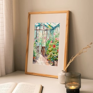 Puede incluir: Una pintura de acuarela enmarcada de un invernadero con un marco de madera. La pintura representa un invernadero lleno de plantas y flores, con una puerta que conduce al exterior. La pintura est&aacute; sobre una mesa blanca con un libro y un portavelas.