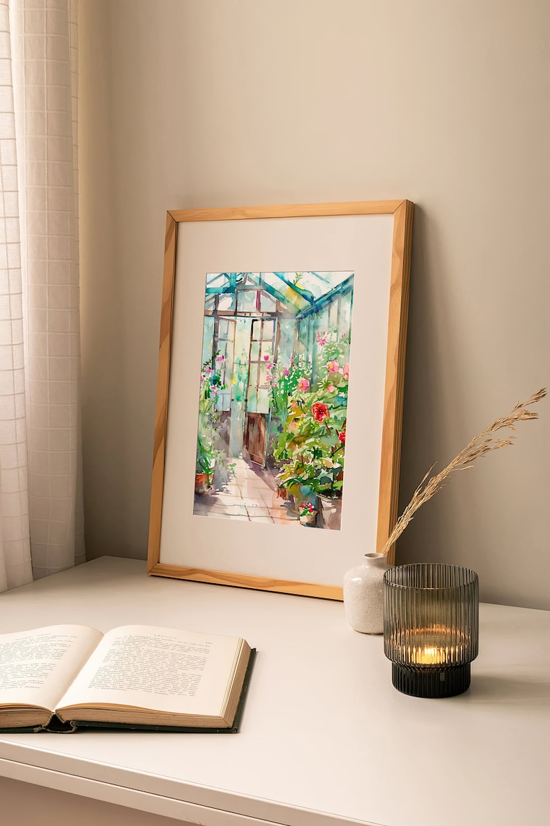 Puede incluir: Una pintura de acuarela enmarcada de un invernadero con un marco de madera. La pintura representa un invernadero lleno de plantas y flores, con una puerta que conduce al exterior. La pintura est&aacute; sobre una mesa blanca con un libro y un portavelas.