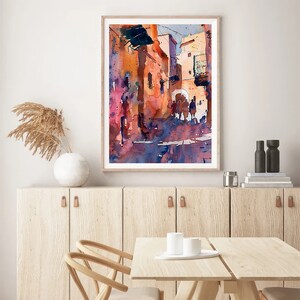 May include: Watercolor painting of a sunlit street scene with buildings in shades of orange, pink, and blue. The artwork is framed in a light wood frame and depicts figures walking along the street. The painting is displayed on a white wall above a wooden cabinet.