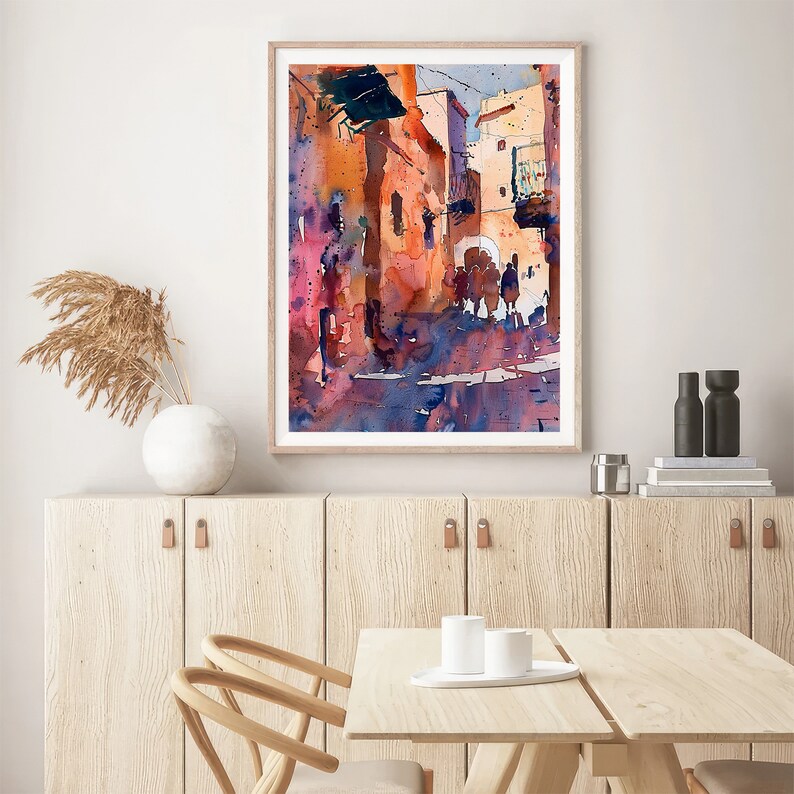May include: Watercolor painting of a sunlit street scene with buildings in shades of orange, pink, and blue. The artwork is framed in a light wood frame and depicts figures walking along the street. The painting is displayed on a white wall above a wooden cabinet.